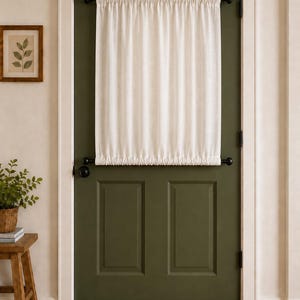 May include: A solid off-white door curtain featuring a gathered fabric design, suspended between two black metal rods mounted horizontally on an olive green door. The curtain covers the upper portion of the door, creating a simple, functional window covering.