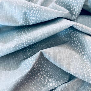 May include: A light blue fabric featuring a subtle white crosshatch grid pattern overlaid with scattered, soft white dots. The material is draped in deep folds, highlighting the textured weave and the contrast between the blue and white elements.