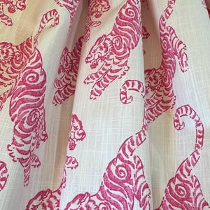 May include: A white fabric featuring a repeating pattern of tigers printed in a vibrant magenta pink. The material displays a visible woven texture with vertical and horizontal threads, and the fabric is draped to create soft, deep folds.
