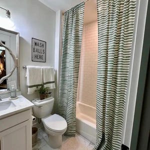 May include: A bathroom with a white toilet, sink, and shower. The shower has a green and white striped curtain. A sign on the wall reads "WASH BALLS HERE". White towels hang on a rack above the toilet.
