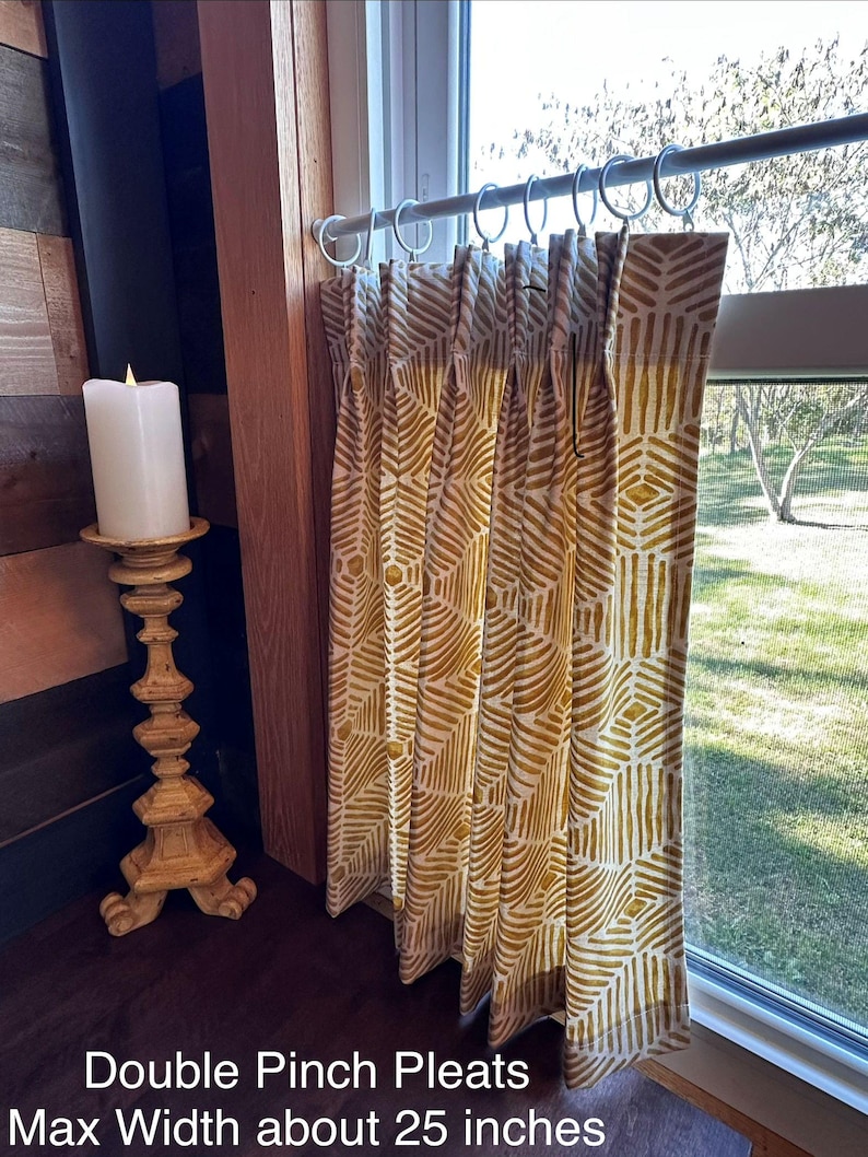 a window with a double pinch pleat curtain hanging from a rod, and a white candle on a wooden stand next to the curtain.
