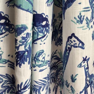May include: A white fabric curtain featuring a repeating pattern of giraffes, palm trees, and tropical foliage in shades of dark blue and light teal. The textile has a visible woven texture and is gathered into vertical folds.