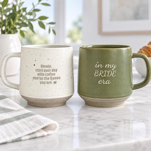 Könnte beinhalten: Zwei Keramikbecher mit Henkeln. Der linke Becher ist cremefarben mit braunen Sprenkeln und dem Text: Nicole, start your day with coffee and be the Queen you are. Der rechte Becher ist olivgrün mit dem Text: in my BRIDE era.