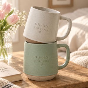 Puede incluir: Dos tazas de cerámica apiladas con acabados moteados. La taza superior es de color blanco roto con el texto "Olivia's grandma" y un corazón. La taza inferior es de color verde salvia con el texto "Olivia's grandpa" y un corazón. Ambas tienen asa.