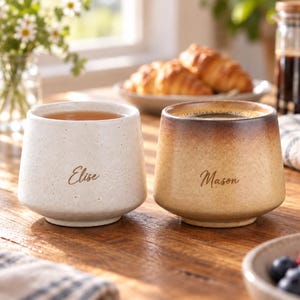 May include: Two ceramic mugs with a tapered shape and speckled finish. The left mug is off-white with the name Elise in cursive script. The right mug features a brown gradient finish with the name Mason in cursive script. Both mugs are handleless.