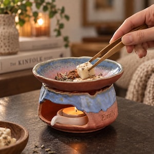 May include: A ceramic wax warmer featuring a two-tone blue and terracotta glaze with the name Emily engraved on the side. The warmer has a wide bowl top for wax melts and a lower opening containing a small white tea light candle holder.