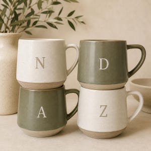 Pode incluir: Quatro canecas de cerâmica dispostas numa pilha de dois por dois. Duas canecas são de cor esbranquiçada com texturas salpicadas e duas são de cor verde sálvia. Cada caneca apresenta uma letra em relevo na frente: N, D, A e Z.