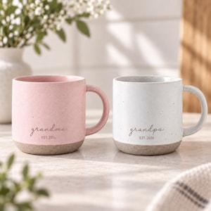 Op de afbeelding: Een roze gespikkelde keramische mok met de tekst grandma EST. 2026 en een witte gespikkelde keramische mok met de tekst grandpa EST. 2026. Beide mokken hebben een gladde bovenkant en een textuurrijke, ongeglazuurde beige onderkant met een oor.