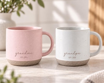 Può includere: Due tazze in ceramica con finitura maculata e base testurizzata. La tazza rosa riporta la scritta "grandma EST. 2026" in caratteri scuri. La tazza bianca riporta la scritta "grandpa EST. 2026" in caratteri scuri. Entrambe hanno un manico.