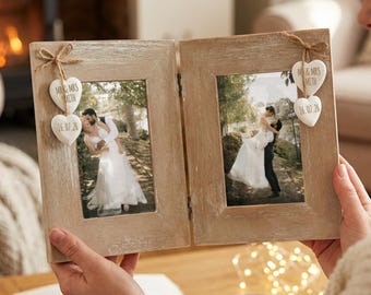Puede incluir: Un marco de fotos doble de madera clara con bisagras y dos aberturas verticales. Cada lado está decorado con dos pequeños adornos blancos en forma de corazón atados con cordel, con el texto "MR & MRS SMITH" y "16.07.26" en tinta negra.