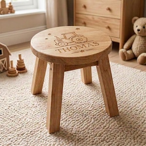 May include: A round wooden stool with four legs featuring an engraved tractor graphic and the name THOMAS on the seat surface. The light-coloured wood grain is visible across the top and legs of this small piece of furniture for children.