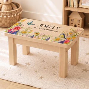 May include: A rectangular wooden step stool with four legs and a flat top surface. The top features a colorful floral print, a blue butterfly, a small bird, and the printed text "EMILY Our Little Wildflower" in a black sans-serif font.