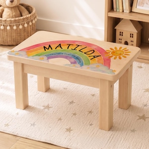 Könnte beinhalten: Ein Hocker aus hellem Holz mit einer bunten Regenbogen-Grafik auf der Oberseite. Das Design zeigt den Namen Matilda in schwarzer Schrift, den Text Somewhere Over the Rainbow sowie eine lächelnde gelbe Sonne mit Wolken und Sternen.