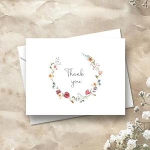 K&ouml;nnte beinhalten: Eine wei&szlig;e, gefaltete Gru&szlig;karte mit einem kreisf&ouml;rmigen Kranz aus Aquarell-Wildblumen in Rosa-, Gelb- und Blaut&ouml;nen. Der Text Thank you ist mittig in einer dunkelgrauen Schreibschrift gedruckt. Auf der linken Seite ist eine kleine Biene zu sehen.