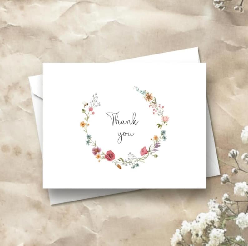 K&ouml;nnte beinhalten: Eine wei&szlig;e, gefaltete Gru&szlig;karte mit einem kreisf&ouml;rmigen Kranz aus Aquarell-Wildblumen in Rosa-, Gelb- und Blaut&ouml;nen. Der Text Thank you ist mittig in einer dunkelgrauen Schreibschrift gedruckt. Auf der linken Seite ist eine kleine Biene zu sehen.