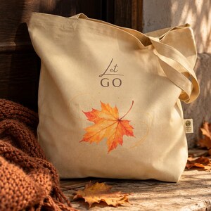 Könnte beinhalten: Eine beige Tragetasche aus Segeltuch mit dem Schriftzug "Let GO" in dunklen Buchstaben über einer Grafik eines leuchtend orangefarbenen und gelben Ahornblatts. Die Tasche hat ein kleines seitliches Etikett und zwei Tragegriffe aus Stoff.
