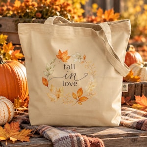Könnte beinhalten: Eine beigefarbene Stofftasche mit einem aufgedruckten Kranz aus herbstlichen Aquarellblättern in Orange-, Gelb- und Brauntönen. Der Text "fall in love" ist mittig in einer dunklen Schreibschrift im Kranz platziert, inklusive kleinem Herz.