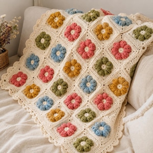 Peut inclure: Une couverture en crochet de couleur crème présentant un motif répétitif de carrés, chacun contenant une fleur en relief à plusieurs pétales dans des tons de rose, jaune, bleu et vert olive. La couverture a un bord festonné décoratif.
