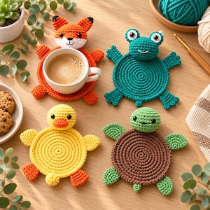 Peut inclure: Quatre dessous de verre en crochet faits main en forme d'animaux : un renard orange, une grenouille sarcelle, un canard jaune et une tortue marron. Le dessous de verre renard porte une tasse en céramique blanche remplie de café.