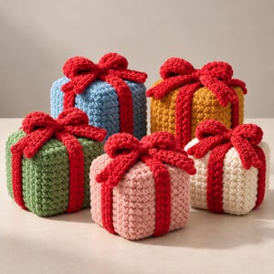 Peut inclure: Un ensemble de cinq boîtes cadeaux carrées au crochet, déclinées en bleu clair, jaune moutarde, vert sauge, rose pâle et crème. Chaque boîte présente un ruban rouge épais et texturé, noué en un grand nœud sur le dessus.