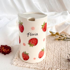 May include: A cylindrical ceramic container with a cream-colored base, decorated with hand-painted red strawberries and small pink polka dots. The name Floria is written in black cursive script on the front. It features a notched top opening.
