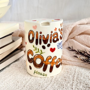 May include: A cylindrical white ceramic mug featuring hand-painted brown lettering that reads Olivia's Coffee please. The design includes small colorful hearts, a green leafy sprig, and coffee bean illustrations scattered across the surface.