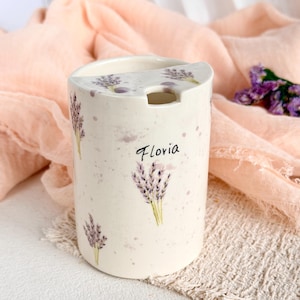 May include: A cylindrical ceramic container with a cream-colored speckled finish, decorated with purple lavender sprigs and the name Floria printed in black script. The top features a semi-circular lid with a small cutout opening for a spoon.