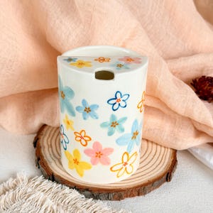 Op de afbeelding: Een cilindrische witte keramische houder met een handgeschilderd patroon van kleine bloemen in tinten geel, lichtblauw, oranje en roze. De bovenkant van de houder heeft een platte rand met een kleine halfronde opening.