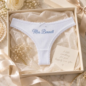 May include: A white ribbed thong featuring a small center bow, lace trim, and the blue embroidered text Mrs. Bennett. The garment is presented in a gift box with tissue paper and a card that reads A little something for your happily ever after.