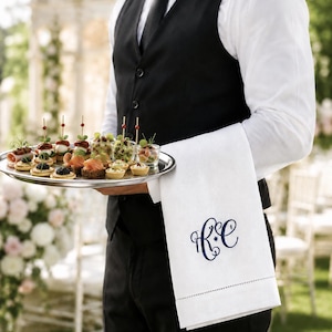 May include: A white linen towel featuring a navy blue embroidered monogram with the letters RC, draped over an arm. The towel has a decorative hemstitch detail near the bottom edge and is held alongside a silver tray of assorted appetizers.