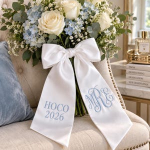 May include: A bouquet of white roses, light blue hydrangeas, and baby's breath tied with a white satin ribbon. The ribbon features light blue embroidery with the text HOCO 2026 on one tail and a decorative three-letter monogram on the other.