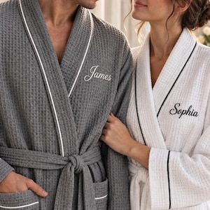May include: A gray waffle-knit robe with white piping and the embroidered name James, alongside a white waffle-knit robe with black piping and the embroidered name Sophia. Both robes feature a shawl collar and a matching tie belt.