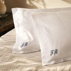 Peut inclure: Deux taies d'oreiller rectangulaires d'un blanc éclatant avec un large bord à volant. Chaque taie est personnalisée avec des initiales brodées en bleu foncé dans une écriture cursive, l'une affichant les lettres JB et l'autre les lettres FR.