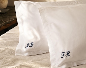 Può includere: Due federe rettangolari bianche con un bordo largo e iniziali ricamate in blu navy. Una federa mostra le lettere JB, l'altra le lettere FR, entrambe cucite con un carattere in corsivo.