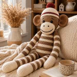 Puede incluir: Un muñeco de mono tejido a ganchillo con rayas horizontales marrones y crema en el cuerpo y las extremidades. El mono lleva un gorro color crema con un pompón rojo oscuro y una raya roja a juego. Tiene orejas grandes y ojos de botón.
