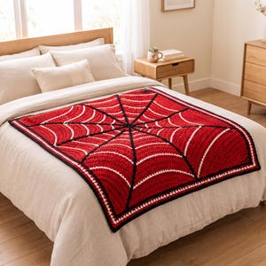 May include: A square crochet blanket featuring a vibrant red spiderweb pattern with thick black web lines and white accent stitching. The blanket has a solid black border and is displayed flat on a neutral-colored bed with pillows.