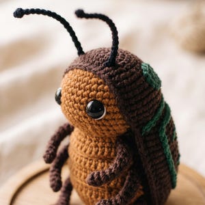 Puede incluir: Un juguete de peluche tejido a ganchillo con forma de escarabajo, con cara y vientre color canela, cabeza y alas de color marrón oscuro y dos largas antenas negras. El escarabajo tiene grandes ojos de plástico negro y detalles verdes.
