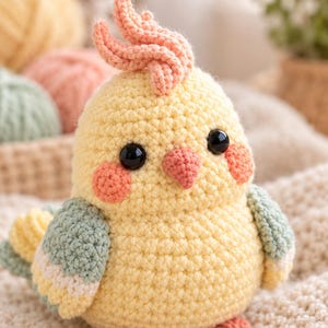 May include: A crocheted cockatiel bird plush toy featuring a pale yellow body, a peach-colored crest, a small peach beak, and round peach cheeks. The wings are detailed with sage green, white, and yellow yarn, and the bird has black eyes.