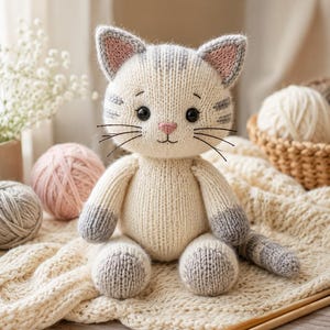 May include: A knitted cat toy featuring a cream-colored body with grey striped markings on the head and tail. The cat has dark embroidered eyes, a pink nose, and grey-tipped paws. The ears are pointed with pink inner details and grey edges.