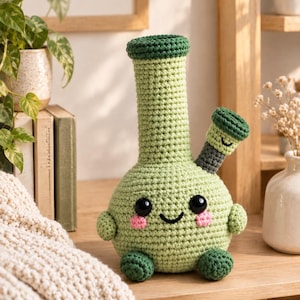 May include: A light green crochet plush toy shaped like a water pipe, featuring a tall cylindrical neck, a round base, and a small side attachment. It has large black plastic eyes, a stitched black smile, and pink embroidered cheeks.