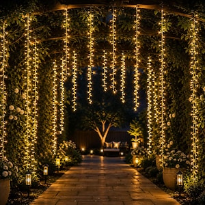 May include: A series of vertical string lights with warm white LED bulbs hanging from an arched trellis. The lights create a glowing curtain effect over a stone pathway flanked by potted plants and small ground-level lanterns in a garden setting.