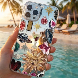 May include: A clear smartphone case featuring various beaded and sequined appliques including a large golden sun with a face, a cocktail glass, a planet with rings, a whale, a fish, a pomegranate, a fig, stars, and seashells scattered throughout.
