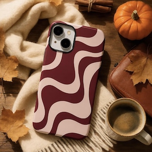 May include: A smartphone case featuring a wavy, abstract pattern in deep burgundy and soft cream. The case has a rectangular shape with rounded corners and a cutout for a dual-lens camera system located in the upper left corner of the back panel.