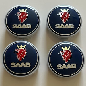 May include: Four circular Saab wheel center caps featuring a dark blue background with a silver border. Each cap displays a red griffin head emblem wearing a gold crown above the silver text SAAB, all finished with a glossy, smooth surface.