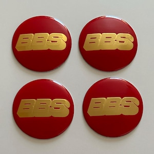 Könnte beinhalten: Vier kreisförmige Radnabendeckel mit glänzend roter Oberfläche und dem goldenen BBS-Logo in der Mitte. Jeder Deckel hat eine glatte, reflektierende Oberfläche, auf der der Text BBS in einer fetten, dreidimensionalen Goldschrift zu sehen ist.