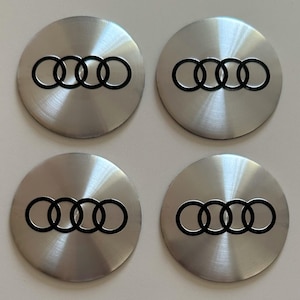 Puede incluir: Cuatro tapacubos circulares de metal plateado, cada uno con un acabado metálico cepillado y un logotipo de Audi negro en relieve que consiste en cuatro anillos entrelazados centrados en la superficie de cada tapa.