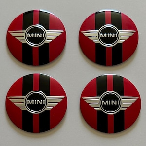 May include: A set of four circular wheel center caps featuring a red and black vertical striped pattern. Each cap displays a central silver MINI logo with stylized wings. The design is glossy and symmetrical, with the word MINI clearly visible.