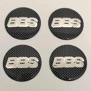 May include: Four circular wheel centre cap emblems featuring a black and grey carbon fibre pattern background. Each emblem displays the silver-coloured text BBS in a bold, raised font centred across the middle of the circular surface.