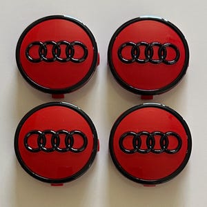 Może przedstawiać: Zestaw czterech okrągłych dekielków do felg z błyszczącym czerwonym tłem i wypukłym czarnym logo czterech pierścieni Audi. Każdy dekielek ma czarną zewnętrzną obwódkę oraz gładkie, odblaskowe wykończenie, ułożone w układzie dwa na dwa.