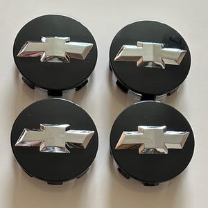 Könnte beinhalten: Ein Set aus vier runden Radnabendeckeln mit glänzend schwarzer Oberfläche und einem markanten, erhabenen, verchromten Chevrolet-Bowtie-Emblem in der Mitte jedes Teils. Diese Autozubehörteile zeichnen sich durch eine glatte, reflektierende Oberfläche aus.